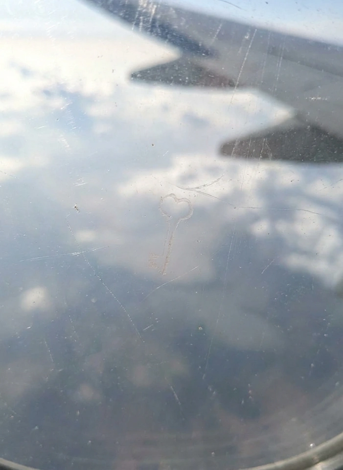 A small key shape in the reflected sky and wing seen through an aircraft window.